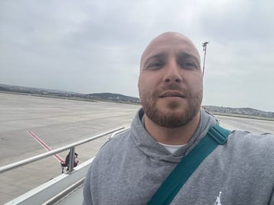 A bald man with a beard stands on an aircraft boarding ramp at Tuzla Airport, Turkey, taking a selfie. He wears a grey hooded sweatshirt with a teal shoulder strap. The airport tarmac stretches behind him, marked with red lines. In the distance, a cityscape sits against a grey sky. A weather station is visible in the background to the right of the man's head. A person is seated on a chair near the ramp.