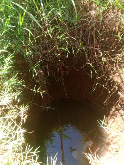 This image depicts a circular excavation in the ground, located in the city of Kiangochi, Kenya. The hole, approximately 1 to 1.5 meters in diameter, features dark brown soil walls with visible root systems and clumps of green grass along its upper rim. The lower portion of the excavation is partially filled with dark, turbid water, reflecting a blue sky and green vegetation. A slender, dark brown stick or branch is partially submerged in the water, positioned vertically from the upper left portion of the water's surface towards the center. No individuals or active interactions are visible within the frame. Sunlight casts shadows on the right side of the excavated soil.