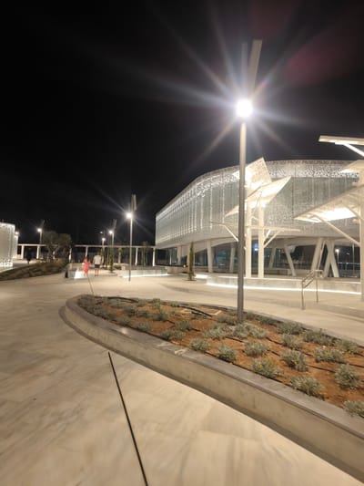 A street-level, eye-level, night-time, long shot shows a modern public area in Paphos Municipality, Cyprus. The area features a paved walkway with a slight curve, bordered by a raised planter filled with low-lying, grey-green vegetation and yellow flowers. In the background, a large, white, modern building with a perforated, geometric facade is illuminated. A streetlamp with a bright, starburst effect illuminates the scene. To the left, two figures walk away from the camera. The overall scene is well-lit and clean, suggesting a contemporary urban environment.