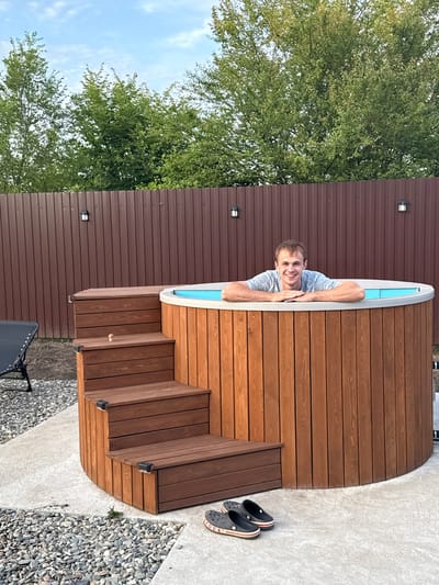 A man with short brown hair and a light blue and grey striped t-shirt smiles as he rests his arms on the edge of a wooden hot tub. He is leaning out of the tub, with his head and upper body visible. The hot tub is circular and made of wooden planks. Steps, also made of wood, lead up to one side of the hot tub. The scene appears to be an outdoor setting, with a brown metal fence in the background and lush green trees. The sky is visible, suggesting it is daytime with some clouds. A pair of black Crocs with red and white stripes are placed on the concrete ground next to the hot tub. There is no visible text in the image. The setting is likely a backyard or a recreational area in Starokorsunskaya, Russia. The man appears happy and relaxed.