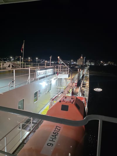 This image captures a nighttime view aboard a ferry docked in the port of Brindisi, Italy. In the foreground, an orange lifeboat is prominently displayed, with the registration number "9 HA6127" clearly visible. The ferry itself is a large vessel, as indicated by the white railing and deck. Further back, a white superstructure of the ferry is visible, bearing a lit sign that reads "EUROCARGO TRIESTE". A flag, partially obscured, flies from a flagpole on the upper deck. The background shows the illuminated coastline of Brindisi, with buildings and port facilities visible under the night sky. The water beside the ferry reflects the lights from the shore and the ship. The overall scene is one of a docked ferry at night, with the city lights of Brindisi serving as a backdrop.