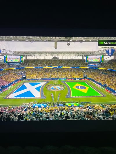 The image shows a wide aerial view of a stadium during a soccer match, likely a pre-game ceremony. The field is prominently decorated with two large national flags: the flag of Scotland on the left and the flag of Brazil on the right. The stadium is filled with a large crowd, predominantly wearing yellow, suggesting support for Brazil. In the background, a large screen displays graphics, and text below it reads "FIFA WORLD CUP 2026." A banner above the crowd on the right side of the stadium displays the text "LiveBall" with "ПРЯМОЙ ЭФИР" written below it, indicating a live broadcast. The overall scene depicts a vibrant and festive atmosphere typical of a major sporting event. The location is identified as the city of Slobozia-Dușca, Moldova, though this specific stadium or event is not visually confirmed by the image itself and would require external information.