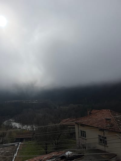 This image captures a landscape in Srednogortsi, Bulgaria, under heavily overcast and foggy conditions. The upper portion of the frame is dominated by a grey, cloudy sky, with a bright, diffused light source, indicative of the sun, visible as a radiant white circle in the upper-left quadrant. A dense band of fog or low-lying clouds obscures the middle ground, transitioning into a dark, heavily forested valley floor.
A river with visible white currents flows through the valley, flanked by numerous deciduous trees, most of which appear bare, suggesting a late autumn or winter setting. In the distant mid-ground on the left, a cluster of buildings or structures with light-colored elements is faintly discernible through the atmospheric haze, situated at the edge of the forested area.
The foreground on the right side features a two-story residential building with light-colored walls and a pitched roof constructed of red clay tiles. Multiple rectangular windows are visible on its facade, and a brick chimney extends from the roofline. Numerous utility wires traverse the scene horizontally across the mid and lower foreground. Behind this primary structure, another building with a similar red-tiled roof is partially visible. Further to the left in the foreground, a smaller, single-story building with a dark red-tiled roof is present, appearing older. The lower-left foreground shows disturbed ground with some green grass and horizontal metal framework, possibly indicating an ongoing construction or development. No individuals or specific actions are observed within the scene.