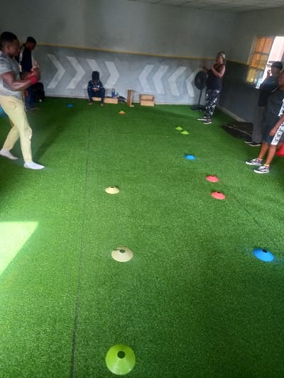 The image shows an indoor space with artificial green turf covering the floor. There are five people present. A man wearing a white shirt and beige pants is facing the camera while holding his left hand to his chest. He is standing to the left of the image. A second man is wearing a black shirt and black pants and is standing to the left of the first man. He is looking to his right. A third man is seated on the floor with his back to the camera. He is wearing a black jacket and black pants. A fourth person is standing on the right side of the image. She is wearing black pants, a white shirt, and a white hat with a black band. Her hands are raised to her shoulders. She is looking to her right. A fifth person is standing to the right of the fourth person. She is wearing black pants and a black shirt with white stripes. Her hands are at her sides and she is looking to her right. Several colored cones are placed at intervals in a line across the artificial turf. The location is in Ita Oniyan, Nigeria, based on coordinates (latitude: 7.294, longitude: 5.157). The overall impression of the scene is one of a sports training exercise taking place indoors.