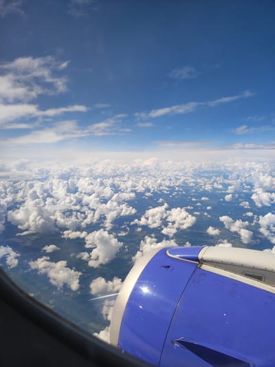 This image is an aerial view from an airplane window, showing a landscape dotted with fluffy white clouds. The foreground features the blue and silver exterior of an airplane engine. The landscape below appears to be rural, with patches of green fields and dark forested areas, interspersed with some human settlements. There are no people visible in the image. The bright blue sky above is partly covered with scattered white clouds. The image does not provide any specific details or context that would allow for a precise identification of the location within the city of Vetka, Belarus.
