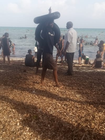 The image depicts a coastal recreational area in Shanzu ward, Kenya, under a clear daylight sky. The foreground comprises a dense layer of desiccated brown marine vegetation covering the sand. A barefoot male, estimated age 18-30, wearing a black t-shirt with "Grapa" text and dark shorts, is centrally positioned, walking towards the right. He carries two black inflatable inner tubes on his right shoulder, one stacked horizontally atop the other, with his right arm extended upwards. Adjacent to him, on his right, an adult male, estimated age 40-60, dressed in a light-colored checkered button-up shirt and dark trousers, stands facing left. To the left of the central figure, a male, estimated age 15-25, in dark attire, sits on the sand with his back to the viewer.
The midground and background feature an estimated 30-50 individuals of varying ages, predominantly adolescents and young adults, engaged in recreational activities. Many are wading or standing in the clear, light blue-green ocean water close to the shore. Others are further offshore, with some utilizing black inflatable inner tubes. The horizon line shows the open ocean under a light blue sky with sparse white clouds.