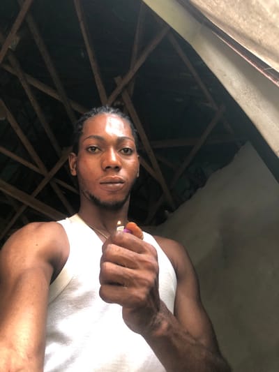 A male subject, approximately 20-35 years old with dark skin, braided hair, and a light beard, is depicted in a waist-up, direct-to-camera portrait. He wears a white ribbed tank top and a thin gold-colored chain around his neck. His right hand is raised, holding an orange and pink plastic lighter that is ignited, displaying a small flame. The background features dark wooden rafters arranged in a crisscross pattern above him, suggesting an exposed ceiling structure. To his right, a section of an unfinished or damaged light-colored wall or partition is visible. The lighting is ambient, with a notable highlight on the subject's forehead. This scene is located in Oraukwu, Nigeria.