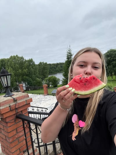 A young woman with blonde hair, wearing a black t-shirt with a cartoon mouse and heart graphic, takes a selfie while holding a slice of watermelon to her mouth. She is on an outdoor balcony overlooking a body of water and a lush green forest. A brick pillar with a black lantern stands to her left, and a metal railing separates the balcony from the landscape. The sky is overcast and cloudy. The location is Zavod Mikhailovskii, Russia.