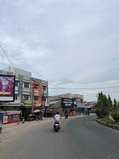 This image captures a street scene in Bundar, Indonesia, on an overcast day. The perspective is a low-angle shot taken from a street level, looking slightly upwards. The foreground is dominated by a paved road, with a single motorcycle rider in the center, wearing a white shirt with red accents and dark pants. They are facing away from the camera, moving along the road.
To the right of the road, a row of multi-story buildings lines the street. The buildings exhibit a mix of architectural styles and colors, with some featuring modern facades and others showing signs of age. One building prominently displays a black banner with white text that reads "TOKO BANGUNAN PANDA PRIMA KERAMIK." Another building, painted in vibrant orange and gray, has multiple windows and air conditioning units. Below these buildings, several small shops and kiosks are visible, some with colorful awnings. A large billboard advertising "GRAPE" and "35.000/POD" is partially visible at the bottom left.
On the left side of the road, a grassy embankment slopes upwards, with trees and power lines creating a slightly cluttered urban landscape. Several other motorcycles are parked or in motion further down the street, indicating a typical day in a populated area. The sky is cloudy, suggesting either early morning or late afternoon, or simply a day with overcast weather. The overall impression is one of a busy, functional street in an Indonesian town.