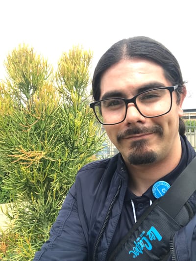 A male individual with dark, shoulder-length hair and black-framed rectangular spectacles is depicted in a close-up, self-portrait-style photograph. He has a mustache connected to a goatee and exhibits bilateral dimples with a slight smile. The subject wears a dark blue zippered jacket over a dark blue collared shirt, to which a circular blue pin displaying partial text "your future" and a white logo resembling a hand is affixed. A black shoulder bag strap bearing a light blue "Lee Cooper" logo crosses his chest. To the left and behind the subject is a prominent *Euphorbia tirucalli* (pencil cactus) plant with numerous thin, green, and yellowish-orange branches. The background consists of an overcast white sky, with faint outlines of modern buildings and a metal fence visible behind the plant and the subject's right shoulder. A light-colored ground surface is partially visible at the lower left. The scene is located in Midrand, South Africa.