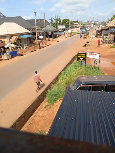 The image depicts a street scene in Achalla, Nigeria, under a partly cloudy sky. A paved road runs through the center, lined with shops and buildings on both sides. The shops have corrugated metal roofs and open fronts displaying goods. Several people are visible, including a lone individual walking on the sidewalk, a person in a red shirt walking on the road, and others tending to stalls. Vehicles such as a truck, a motorcycle, and a tricycle are present on the road. A sign indicating "COTTAGE GUEST HOUSE BAR RESTAURANT ACCOMMODATION" and another for "CHIDIOMIMI HOSPITAL" are visible. The ground is a reddish-brown earth, and there is a drainage ditch running alongside the sidewalk. A black vehicle with a corrugated metal roof partially covers the foreground of the image.