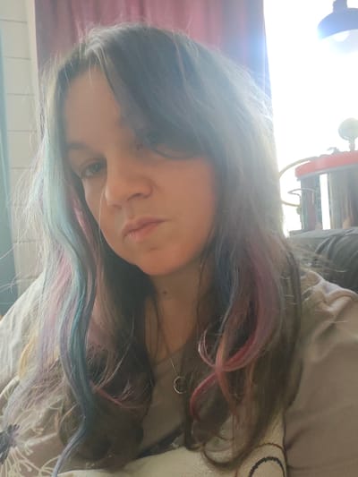A woman with long, dark hair streaked with blue and pink highlights is photographed indoors. She wears a light gray t-shirt and a necklace with a circular pendant. Her expression is neutral. A lamp is visible in the background, partially obscured by bright sunlight. The location in Свистуха (кузяевский С/о), Russia cannot be verified from the provided image.