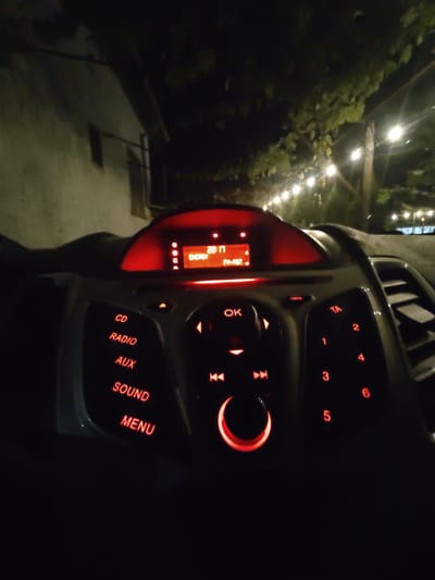 The image shows the interior of a car at night, specifically the dashboard and center console of a Ford Fiesta. The car radio display is illuminated with red light, showing "22:17" and "ENERGY FM-RST". Buttons with red lettering are visible, including "CD", "RADIO", "AUX", "SOUND", "MENU", "OK", and numerical buttons "1" through "6". A rotary dial with a red illuminated ring is also present. Outside the car, string lights are visible in the background, illuminating trees and the facade of a building. The setting appears to be an outdoor area, possibly a restaurant or a residential property, at night. There are no people or specific actions depicted in the image. The location is identified as Tervel, Bulgaria.