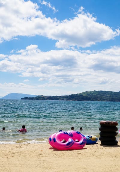 This image captures a serene beach scene at Lumban Bul Bul, Indonesia, under a bright blue sky dotted with fluffy white clouds. The foreground is dominated by a sandy beach with several inflatable tubes, one a vibrant pink with purple accents, and another a blue and yellow tube. To the right, a stack of black inner tubes stands tall. In the shallow, turquoise water, a few children are visible, some playing and wading, while others are partially submerged. The background features the expansive, shimmering water of a lake or sea, with a lush green, mountainous coastline stretching across the horizon. The overall atmosphere is one of peaceful recreation and natural beauty.