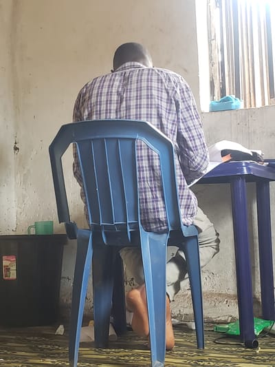 In Kachia, Nigeria, a person is seated at a blue plastic table, facing away from the camera, with their head bowed as if engaged in an activity like studying or writing. They are wearing a checkered long-sleeved shirt and light-colored trousers. The person is seated on a blue plastic chair. The table is covered with papers and what appears to be a white cloth. In the background, a window with vertical bars allows sunlight to stream in, illuminating the scene. To the left of the table, a black plastic container with a label is visible, and a green mug sits on top of it. The environment appears to be a simple room with a concrete floor and textured walls.