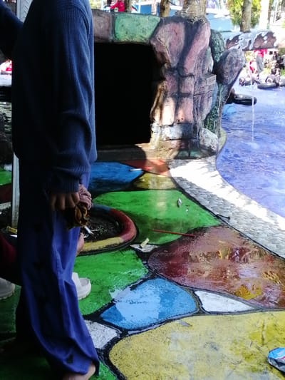 The image depicts an outdoor scene with a pool and a person standing in the foreground. The person is wearing a dark blue long-sleeved shirt and blue pants, holding a piece of cloth in their right hand. The ground around the pool is painted in a colorful, mosaic-like pattern of green, blue, yellow, and red. To the left of the person is a dark opening, possibly an entrance, with a rocky, artificial structure above it. In the background, to the right, is a pool of blue water where several people are visible floating on inner tubes. There are also trees and a structure with a white and brown striped roof in the distance. The overall impression is that of a public pool or water park.