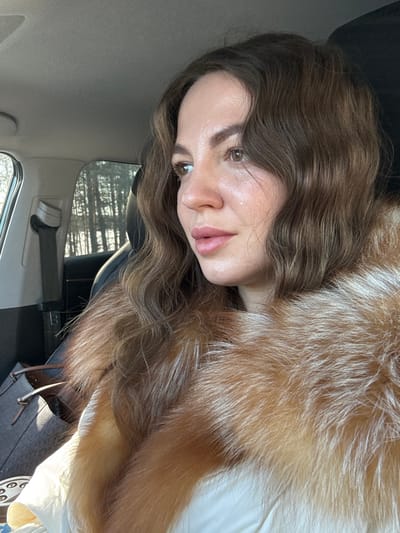 A woman is seated in the passenger seat of a vehicle in the city of Nazhitsy, Russia. She is facing towards the left side of the frame, looking out the car window. The woman has medium-brown wavy hair and brown eyes. She is wearing a light-colored outer garment with a prominent reddish-brown fur collar. A tear is visible on her left cheek, below her eye. The car's interior features a light-colored ceiling, a dark headrest behind her, and a gray seatbelt on the left. A dark bag with light-colored straps rests on the seat next to her. Through the window, a sparse forest of trees is visible under a bright sky, suggesting a daytime setting.