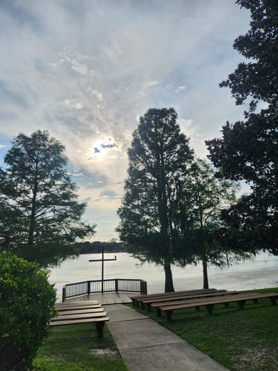 This is a full shot, eye-level, outdoor photograph of an outdoor chapel setup next to a body of water, likely Lake Sybelia in Maitland, Florida. In the foreground, two sets of empty wooden benches are positioned on a grassy area to the right of a concrete walkway. The walkway leads towards a wooden deck with a railing, where a large, dark cross stands as the focal point. Beyond the deck is the calm, shimmering surface of the lake, reflecting the bright sky. Several tall, green trees with wispy foliage frame the scene on the left and right, with the sun peeking through a partly cloudy sky, creating a serene and tranquil atmosphere. The overall impression is one of peace and natural beauty.