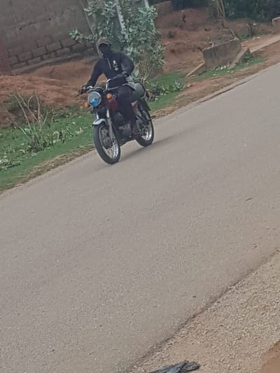 A motorcyclist is on a paved road in what appears to be Hanwa, Nigeria. The rider is wearing dark clothing and a helmet. The motorcycle is parked near the side of the road, with vegetation and a brick structure nearby. A dirt or gravel shoulder is visible to the right of the pavement.