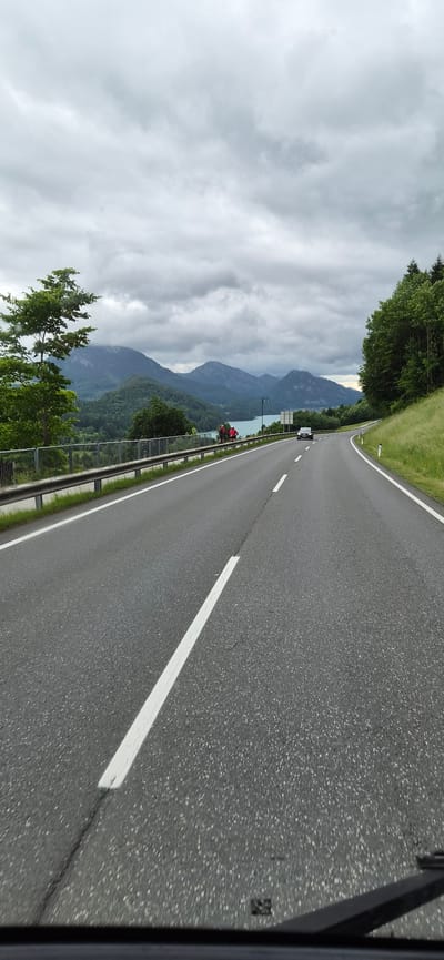 The image shows a road in Hof bei Salzburg, Austria, with a mountainous landscape in the background. The road is asphalt and has white lines marking the lanes. There is a guardrail on the left side of the road, with grass and trees behind it. In the distance, there are mountains and a lake. The sky is cloudy. There are two people on bicycles on the side of the road, and a car is driving on the road. The image is taken from a moving vehicle.