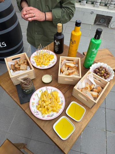 An outdoor tasting event is depicted on a light brown wooden table, featuring various olive oil products and accompanying food items, set in Felgueiras, Portugal.
One adult male is partially visible on the left, from the waist up, wearing a dark green long-sleeved shirt and light-colored trousers. His hands are clasped at waist level, displaying a silver ring on his left ring finger and a dark beaded bracelet on his left wrist.
The table holds three distinct olive oil bottles from the "Alma do Bago" brand: a black bottle with a gold cap ("PICUAL"), a yellow bottle with a black cap ("ARBEQUINA"), and a green bottle with a black cap ("BIOLOGICA").
Food items include:
* Two white, floral-patterned plates, each laden with yellow pineapple cubes, individually pierced with wooden toothpicks.
* Three small wooden crates lined with white paper. The leftmost crate contains small rectangular toast pieces. The other two crates contain slices of baguette-style bread.
* A white, fluted, flower-shaped bowl filled with a mix of green and black olives.
* A small glass jar of dark, textured olive tapenade ("Alma do Bago" branded), accompanied by a small wooden spoon/spreader, resting on a small dark slate board.
* Two square, white ceramic dishes, each containing golden-yellow olive oil.
* A round, silver-rimmed lid is placed open on the table.
The background shows a light gray tiled ground. To the far left, a black wooden barrel with partial white lettering ("ES") is visible. Behind the table, a white-painted building facade with a dark railing and a ground-level grate is present. The scene is arranged for sampling and presentation.