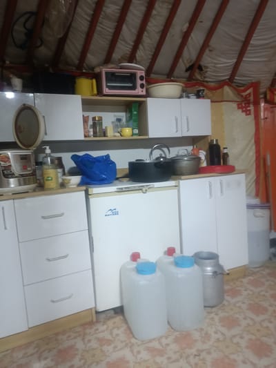 The image depicts the interior of a ger, a traditional Mongolian dwelling, likely located in Ulaanbaatar, Mongolia, given the context. The scene is a kitchen area within the ger, characterized by the distinctive conical roof structure supported by wooden poles, covered with white fabric.
The kitchen setup includes white modular cabinets and drawers. On the top shelf of a cabinet, there is a pink mini oven, a yellow kettle, and a white bowl. Below this shelf, a rice cooker is visible on the countertop, alongside a bottle of liquid soap and some jars. A blue plastic bag rests on the counter, and behind it, a black cooking pot and a smaller metal pot with a lid are present. To the right of the pots, there is a red circular tray with several bottles on it.
In the center of the image, a white refrigerator with a "MATS" logo is positioned. In front of the refrigerator, there are three large white plastic containers with blue lids, and a silver milk churn. The floor is covered with a patterned carpet. The overall impression is one of a functional, albeit somewhat cluttered, domestic space adapted for use in a traditional dwelling.