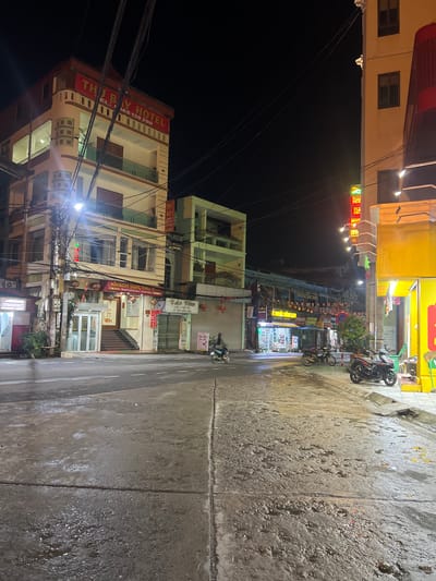 The image depicts a street scene at night in Bắc Hà, Vietnam. A multi-story building with the prominent sign "THU BAY HOTEL" and "SATURDAY HOTEL" is visible on the left. The building is illuminated by exterior lights and has a beige and red facade with balconies. Adjacent to the hotel are various businesses with illuminated signs, including "AUTOBANK" and "KHACH SAN THU BAY." The street in the foreground appears wet and reflective, suggesting recent rain. Several motorcycles are parked along the sidewalk. In the background, other buildings with lit signs and awnings line the street, indicating a commercial area. Red lanterns are suspended along some of the storefronts. The sky is dark, with overhead wires crisscrossing the frame. The overall impression is of a lively, illuminated street scene at night in a Vietnamese town.