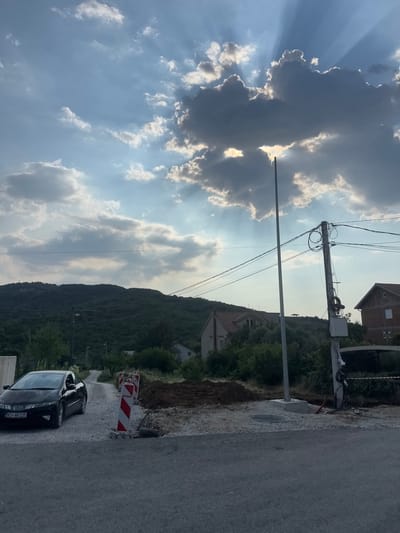 A person is working on a utility pole against a backdrop of a cloudy sky with sunbeams. The scene depicts an outdoor environment, likely a residential area given the houses visible. The utility pole is adorned with numerous wires, suggesting an electrical or telecommunications infrastructure. The ground is a mix of gravel and dirt, with some construction barriers indicating ongoing work or road repair. A black car is parked on the asphalt road. The time of day appears to be late afternoon or early evening, judging by the quality of the light and the position of the sun. The weather seems partly cloudy, with dramatic shafts of sunlight breaking through the clouds. The overall impression is one of active work in a somewhat rural or suburban setting, possibly in Radanovići, Montenegro. No legible text is visible in the image.