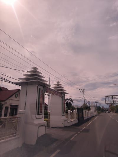 This image captures an exterior view of an ornate gateway structure on the left side of a paved road, likely in Lampeudeu Baroh, Indonesia. The gateway consists of two prominent white pillars, each topped with multiple stacked, rectangular, roof-like tiers. These pillars are adorned with green trim along their bases and vertical edges.

A banner on the right pillar prominently displays the text "SELAMAT HARI RAYA Idul Adha" followed by "1444 H / 2023 M", indicating the celebration of Eid al-Adha in the year 2023.

To the immediate left of the gateway, part of a building with a red-tiled roof and a white facade, featuring columns and an emblem, is visible. A white fence with a yellow-gold railing extends from the gateway toward this building.

The sky is cloudy, but a bright sunbeam is visible from the top-left corner, suggesting daytime. Numerous overhead power lines traverse the scene, connecting to utility poles that line the road.

The paved road curves into the distance on the right, with visible lane markings. Several cars and motorcycles can be seen in the distance, indicating traffic. Along the right side of the road, there is a low white fence, and further down, another smaller, similarly styled white structure, along with palm trees and a large metal framework, possibly for a billboard. The scene depicts a typical street environment in a semi-urban or suburban area.