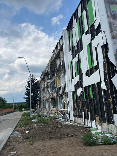 The image displays the heavily damaged exterior of a multi-story building located in Staryi Saltiv, Ukraine. The structure's facade features remnants of white and green cladding, extensively ripped and fragmented, exposing dark underlying insulation and structural supports. Numerous windows are shattered, missing, or severely damaged, with their frames twisted and interiors visible. A significant amount of debris, including shattered glass, metal, and pieces of the building's facade, lies scattered on the dirt ground directly in front of the structure. To the left, a paved sidewalk runs parallel to the building, alongside a street light pole and a tall conifer tree. In the background, a fence and a distant treeline are visible under a partly cloudy sky. The scene is devoid of people or active subjects.