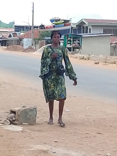 A woman wearing a green dress with a floral pattern walks along a dirt road in Iseyin, Nigeria. She has a dark purse slung across her body. The woman is wearing flip-flops. In the background, there are buildings with corrugated metal roofs, a small shop with shelves stocked with items, and some piles of dirt. A utility pole stands tall in the distance. The sky is overcast, suggesting a cloudy day. The scene appears to be set during daylight hours. The woman's facial expression is not clearly discernible.