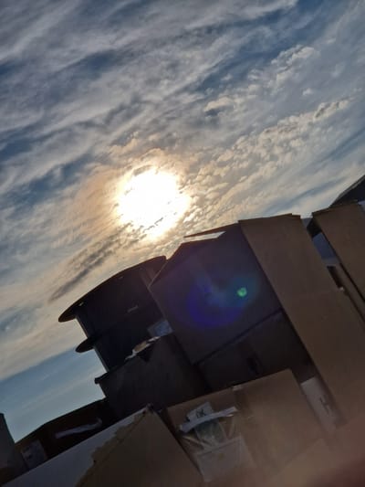 The image shows a sunlit sky with wispy clouds. In the foreground, there are stacks of cardboard boxes. The sun is bright and positioned high in the sky, casting light on the clouds and creating a lens flare in the lower part of the image. The cardboard boxes appear to be in a somewhat disorganized state, with some open and others stacked. There are no people visible in the image. The overall impression is an outdoor scene, possibly an urban or industrial area, given the presence of boxes that might be awaiting delivery or disposal. The tilt of the camera suggests an upward angle of view towards the sky. There are no specific architectural details that would definitively place the location within Bahovitsa, Bulgaria without further context.