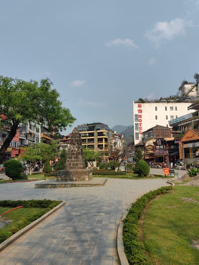This image depicts a public square or park area in the city of Sa Pa, Vietnam, on a clear day with a light blue sky and scattered white clouds. In the foreground and center-left, a weathered dark grey stone monument, constructed from stacked rectangular blocks, stands on a raised platform. Paved grey tiled pathways curve through the scene, bordered by neatly maintained green grass and low green hedges. A large leafy green tree occupies the left foreground, partially obscuring multi-story buildings. A red coiled hose lies on the grass near the left pathway.
Beyond the park, a two-lane paved street is lined with numerous commercial buildings. On the left side of the street, various shops and restaurants are visible, with signage primarily in Vietnamese. A grey SUV is partially visible parked on this side. On the right side of the street, a prominent white multi-story building displays large vertical red text "HAI YEN HOTEL." Below it, other businesses include an "LPBank" branch, "NEW WORLD," and "SAPA HOTPOT BBQ-HOTPOT," with associated signage and parked motorbikes. Several individuals are visible on the sidewalks and near the parked vehicles. A green park bench and a red sign with Vietnamese text "CẤM" (FORBIDDEN) are positioned on the right side of the main road. Distant mountains are visible in the background, behind the urban structures.