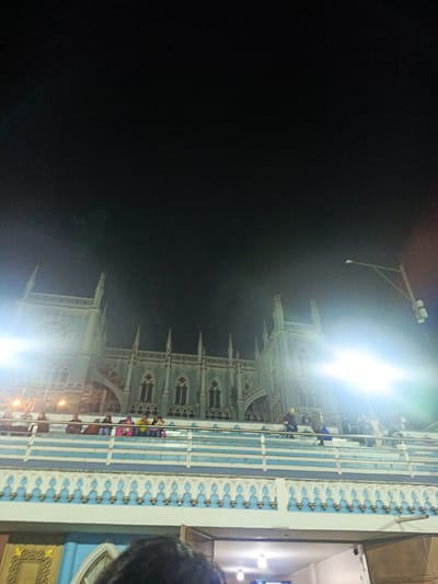 The image shows the Sanctuary of Our Lady of El Cisne at night, located in El Cisne, Ecuador. A section of the building is illuminated by bright lights. People are sitting or standing on a balcony in front of the structure. The sky is dark. A street lamp is visible to the right. A portion of the building's lower level is also visible.