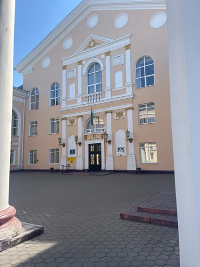 The image shows the exterior of a building, likely a public institution, located in Temirtau, Kazakhstan. The building has a peach-colored facade with white and gold decorative elements, including columns, arched windows, and a prominent central entrance. A flag, likely the flag of Kazakhstan, is visible flying from a pole above the entrance. The foreground features a paved courtyard with hexagonal tiles, leading up to the building. A large white column is present on the left side of the image, partially obscuring the building. The sky is clear and blue. There are no people visible in the image.