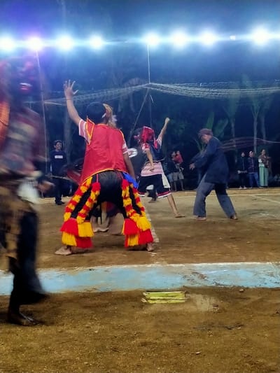 The image depicts an outdoor night performance in Tamansari, Indonesia, illuminated by a string of bright overhead lights against a dark, tree-lined background. The ground is unpaved earth, marked by a light blue and white painted border. In the blurred left foreground, a figure wearing a patterned garment and a red mask is in motion. The central performer, viewed from behind, wears a red vest over a light shirt, dark pants, and black trousers heavily fringed with red and yellow tassels; their right arm is raised, and a dark cap is worn. Adjacent, a smaller performer in a striped shirt, dark shorts, and a red head covering also has a raised right arm. To the right, a barefoot male performer in dark blue attire and a dark cap is in a low, bent stance. In the left background, a smiling male spectator stands near a dark object. A group of approximately five to seven spectators, including adults and a child, observe from the right background. A white net structure is suspended across the upper background. A small, light-colored rectangular object rests on the ground within the painted border in the near foreground.