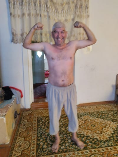 A full-body shot shows a smiling, bald, middle-aged man with light skin, wearing light gray shorts and no shirt. He is standing indoors, flexing his biceps, with his arms bent at the elbows and his fists raised to shoulder height. The man's head is tilted slightly to his right. Behind him, a patterned curtain with a floral design is visible. To the left of the man, a doorway opens into another room with red and patterned furnishings. The man is standing on a patterned rug that covers the wooden floor. A small table with a patterned surface and some folded clothes is partially visible to the far left. The overall lighting is dim, suggesting it is indoors during the day. The image appears to be taken in a residential setting in Yangibazar, Uzbekistan.