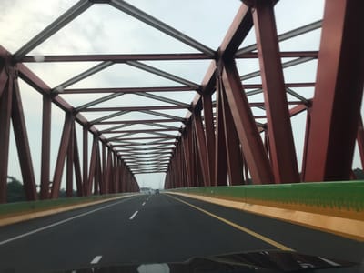 A view from inside a vehicle traveling on a multi-lane highway running through a steel truss bridge. The bridge is painted in a red hue, with exposed steel beams forming a triangular lattice overhead. The highway has painted white and yellow lane markings. The sky is overcast and light grey. There is a green-painted guardrail lining the side of the highway. Other vehicles are visible in the distance on the highway. The vehicle's dashboard is partially visible at the bottom of the frame. The location is Gohor Lama, Indonesia.