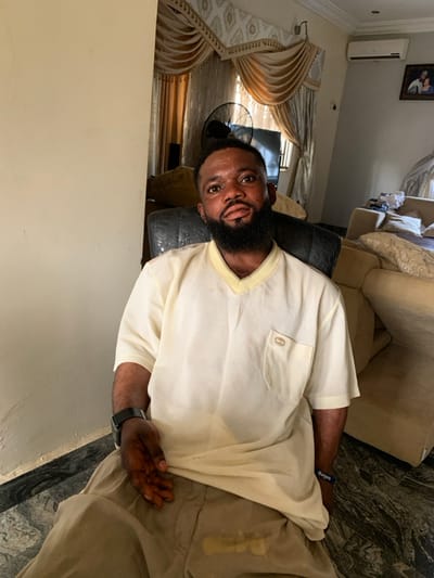 The scene depicts an adult male seated in an indoor living room setting in Eika-Adagu New Layout, Nigeria. The subject, a dark-complexioned male with a full beard and short dark hair, is wearing a light cream V-neck short-sleeved shirt with a left chest pocket featuring a small brown emblem, and light brown trousers. A black smartwatch is on his left wrist, and a blue wristband is on his right. He is seated in a dark, possibly leather, armchair, looking directly at the camera with a neutral expression.
The background shows light-colored walls and a floor tiled with a grey and white marble-like pattern. A light-colored sofa is partially visible to the right. Behind the subject, a window is covered by elaborate light gold/beige curtains with ornate valances. A black standing fan is positioned to the left of the curtains, in front of a partially visible flat-screen television. A wall-mounted air conditioning unit and a framed picture are visible on the wall to the right of the curtains. A light-colored wall partition is present on the left side of the image. The subject is stationary, posing for the photograph.