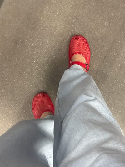 The image shows a person wearing light blue jeans and red mesh ballet flats with a buckle on the strap. The shoes appear to be slightly worn. The person is standing on a textured, grey surface, possibly asphalt or concrete, which has small flecks of white and lighter grey mixed in. The scene is photographed from a high angle, looking down at the person's feet and legs. There are no discernible landmarks or specific architectural features that would allow for precise identification of a location within Ivanovo, Russia. The lighting suggests an outdoor or well-lit indoor environment.