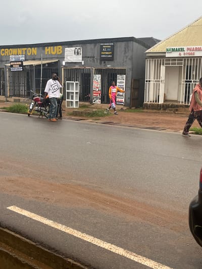 The image depicts a street scene in Sabojibi, Nigeria, with a focus on storefronts and a few individuals. The time appears to be daytime, with overcast skies suggesting possible rain or a generally cloudy day.

The foreground shows a road with a white painted line. On the left side of the road, a building houses several businesses. One prominent sign reads "CROWNTON HUB," with smaller signs below indicating "CHRIS CARE LAUNDRY AND DRY CLEANING SERVICES" and "IMPACT BUSINESS CENTRE" offering printing, photocopying, and coding classes. Another storefront, labeled "MAMAKEL PHARMA & STORE," is visible further down.

Several people are present. A person in a pink and yellow outfit is standing near the "IMPACT BUSINESS CENTRE" signage. Further down, a man in a white shirt with a geometric design and jeans is on a motorcycle, appearing to engage in conversation or look at his phone. Another person in a pink hoodie is walking away from the camera in the background, near the "MAMAKEL PHARMA & STORE."

The overall setting is that of a commercial area, likely a local market or shopping strip. The architecture is typical of the region, with metal security bars on shop fronts. The ground is a mix of asphalt and dirt, with some vegetation visible along the edge of the building. The mood is neutral, with no strong emotional cues evident. The visible text provides specific details about the services offered by the businesses.