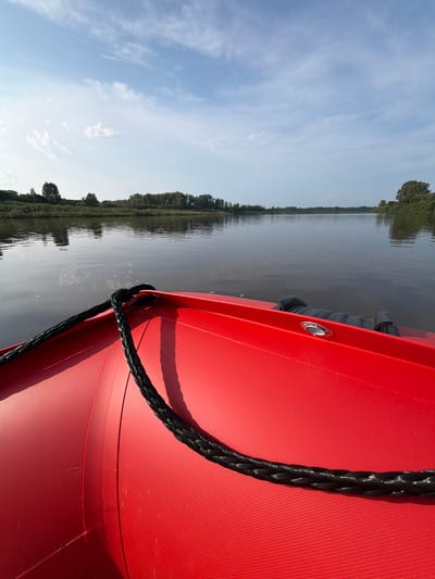 An eye-level, close-up, point-of-view shot from a bright red inflatable boat shows a thick black rope extending across the front of the vessel, secured to an attachment point. The rope is braided and has metallic threads woven into it, reflecting light. The boat's surface is textured and appears to be a synthetic material. In the background, a calm, dark body of water reflects the sky and the distant tree line. The sky is a pale blue with scattered white clouds. The distant shore is lined with dense green trees and foliage. The image appears to be taken from a slow-moving boat on a river or lake, possibly in the city of Kazaevo, Russia, given the landscape.