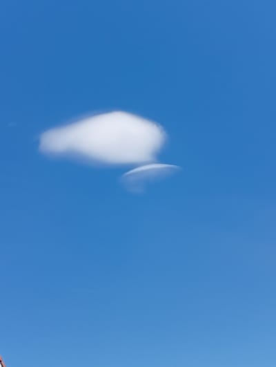 The image shows a clear blue sky with two clouds. One cloud is larger and puffier, located in the upper left portion of the frame. Below and slightly to the right of the larger cloud is a smaller, more elongated cloud. A faint contrail from an aircraft is visible in the far upper right corner. The bottom left of the image shows a sliver of a reddish-brown roof structure, possibly from a building. The scene depicts an outdoor environment with a focus on the sky. The location in Murten, Switzerland, is not discernible from the image itself.