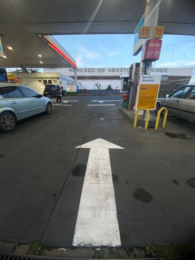 This image shows a gas station called "Mercado de Abasto" in Quilmes, Argentina. A white painted arrow points towards the front of the station. A car is parked near a gas pump, and a person is standing nearby. A yellow sign reads "Servicio Atendido" and includes rules for fueling. The sign also indicates that GNC (compressed natural gas) service is available. The gas station has signs for Shell Helix motor oil.
