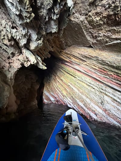 A person is stand-up paddleboarding in a cave. The paddleboard is blue with orange accents and white stripes. It is equipped with a paddle, a foot strap, and a leash. The cave walls are made of rock formations, with some areas exhibiting striations of red, white, and yellow. The water in the cave is dark and appears calm. The overall impression is one of exploration and adventure within a natural geological formation. The lighting suggests an artificial light source is illuminating the cave. No people are clearly visible, but the presence of the paddleboard and gear implies a person is operating it.