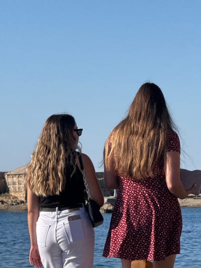 Two women are standing outdoors facing away from the camera, with a body of water in the foreground and a clear blue sky above. The woman on the left wears a black tank top and white jeans, and she has long, wavy blonde hair and is wearing sunglasses. She has a black crossbody bag. The woman on the right wears a red floral print dress and holds a light-colored hat in her right hand. In the background, there is a stone structure partially visible. The scene appears to be a waterfront area, possibly in the city of Chania, Greece, given the architectural style of the background structure and the general appearance of the waterfront.