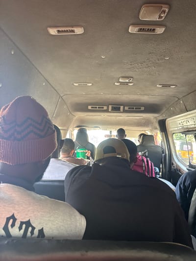 The image depicts the interior of a crowded passenger van, likely a public transport vehicle, somewhere in the city of Orifite, Nigeria. Several individuals are visible seated close together, with a range of clothing and headwear apparent including a rust-colored beanie. The van's interior features air vents along the ceiling. Luggage and other personal items are stored above the seats. The lighting suggests a bright, possibly sunny, day.