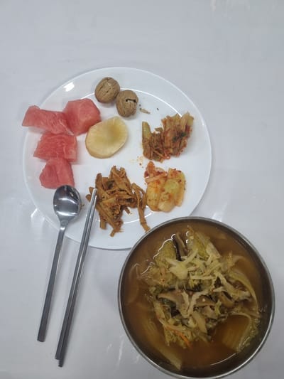 In the city of Mogi das Cruzes, Brazil, a white plate with food is placed on a white surface. On the plate, there are pieces of watermelon, two walnuts, a slice of white radish, and several portions of kimchi. Next to the plate, a spoon and a pair of chopsticks are resting on the white surface. In the foreground, a metal bowl contains a soup with visible napa cabbage and other ingredients. The image appears to be taken from a top-down perspective.