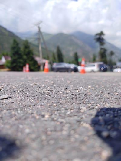 The image is taken at a low angle, close to the asphalt ground. The foreground is in sharp focus, showing the texture of the asphalt with small pebbles. In the midground, several orange traffic cones are visible, blurred due to the shallow depth of field. Beyond the cones, there are out-of-focus vehicles, including a dark-colored car and a white van. In the background, blurry green trees and buildings can be seen, with a power line pole and cables. Further in the distance, hazy mountains dominate the skyline under a cloudy sky. The scene appears to be an outdoor setting, possibly a road or parking area, in a mountainous region. The location is identified as Unknown, Kyrgyzstan.