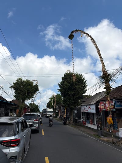 The image depicts a street scene in Tampaksiring, Indonesia, with a narrow road bordered by shops and lush green trees under a bright blue sky dotted with white clouds. Several vehicles, including cars and a motorcycle, are present on the road, suggesting moderate traffic. The shops lining the street display various advertisements and products, with some featuring festive decorations, possibly related to a local celebration. Large, elaborately crafted bamboo and palm leaf structures, known as "penjor," arch over the road, adding to the culturally rich atmosphere. These decorations are characteristic of Balinese traditions. The overall environment suggests a bustling, yet culturally vibrant, urban or semi-urban setting.